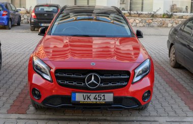 Gaziveren Kıbrıs Rum Kesimi - 30.01.2025 Şık kırmızı lüks bir araba, akşam saatlerinde çağdaş binaların parladığı bir zemin üzerinde duruyor.