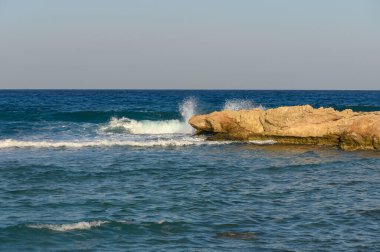 Güneş batarken, yumuşak dalgalar engebeli kayaların üzerinde şakacı bir tur atar ve sakin bir sahil atmosferi oluşturur..