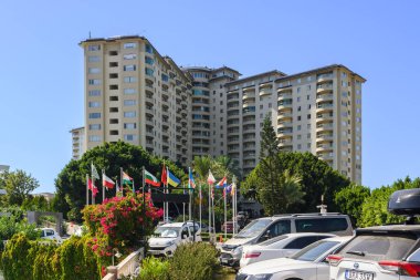 Canlı bir şehir manzarasında parlak güneş ışığı altında renkli bayrakları ve gür yeşilliği olan konut kuleleri yer alır..