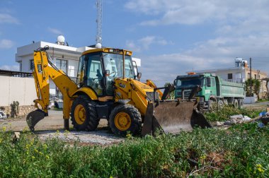 Gaziveren Kıbrıs Rum Kesimi - 03.02.2025 Sarı kepçe yükleyici ve yeşil kamyon yeşil ve binalarla çevrili bir inşaat sahasına park edildi.