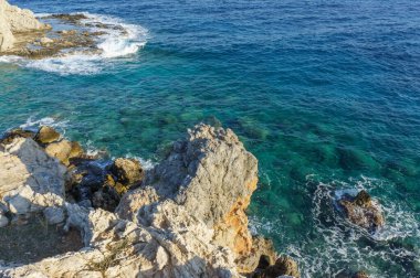 Dalgalar engebeli kayalara hafifçe çarparken, canlı mavi su güneş ışığı altında parlarken, sükunet yaratır..