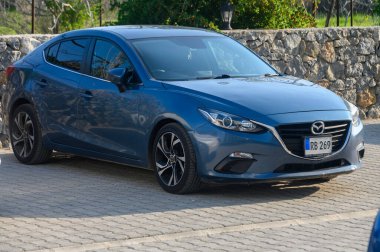 Gaziveren Kıbrıs Rum Kesimi - 16.01.2025 Parlak mavi bir sedan, güneşli bir günün güneşinin altında güneşlenen, resim gibi taştan bir duvarın yanına park edilmiştir..