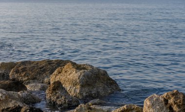 Gün batımında dalgalar taşlara çarpar, dingin yüzeyde ışık parıldar, barışçıl bir hava yaratır..