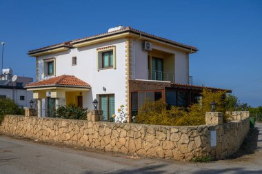 Sessiz bir mahallede güneşli bir günde, taştan bir duvar ve yemyeşil yapraklarla çevrili iki katlı canlı bir ev..