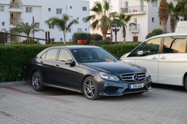 Gaziveren Kıbrıs Rum Kesimi - 30.01.2025 Şık lüks bir sedan, güneş batarken bereketli palmiye ağaçları ve şık binalarla çevrili modern bir arsada dinleniyor.