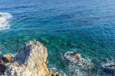 Kristal berrak sular güneşin altında parıldıyor, sakin sahil boyunca engebeli kayaları kucaklıyor..