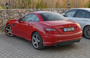 Gaziveren Kıbrıs Rum Kesimi - 30.01.2025 Çarpıcı kırmızı bir üstü açık araba taş bir duvarın yanında duruyor, öğleden sonra güneşinin sıcacık parıltısında güneşleniyor.