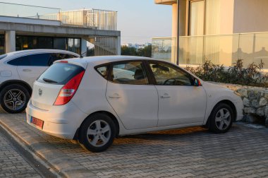 Gaziveren Kıbrıs Rum Kesimi - 30.01.2025 Altın güneş ışığı akşam saatlerinde uzun gölgeler oluştururken, beyaz bir hatchback şık bir binanın yanında dinleniyor..