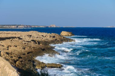 Yumuşak dalgalar dalgalı kıyı şeridine vurur, parlak mavi bir zemin üzerinde güneş ışığını yansıtır..