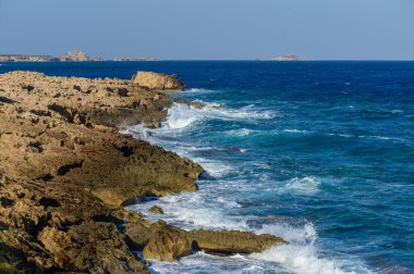 Dalgalar kıyı şeridi boyunca engebeli kayalarla çarpışırken yumuşak dalgalar ritmik bir melodi yaratır..