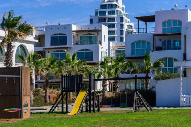 Ekipmanlarla dolu canlı bir oyun alanı berrak mavi gökyüzünün altında, sahil kenarındaki zarif evlerin yanında duruyor..