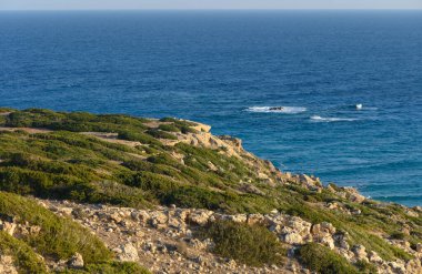 Engebeli kayalıklara bakan güneş sakin denizin üzerinde batarken yumuşak dalgalar kıyıya vurur..