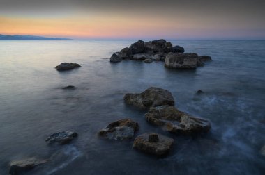 Gökyüzü alacakaranlığa geçerken sakin sulardan kayalar çıkar ve kıyıda huzurlu bir ortam yaratır..