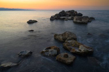 Yumuşak dalgalar engebeli kayalara çarpıyor. Canlı bir günbatımı, kıyı boyunca gökyüzünü altın ve turuncu renklerle boyuyor..