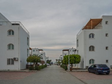 Beyaz binalar ve yeşilliklerle dolu sakin bir patika, kasvetli bir gökyüzünün altında denize doğru ilerliyor..