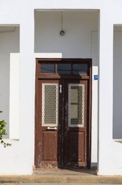 Örgü ile süslenmiş yıpranmış bir kapı güneşli beyaz bir duvarda oturur, sıcak bir günde merakı ve nostaljiyi davet eder..