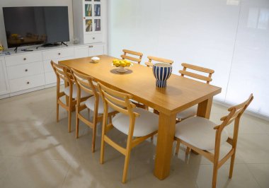 Sleek wooden dining table surrounded by white chairs, showcasing a vibrant citrus bowl in a sunny kitchen environment. clipart