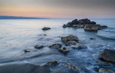 Ufuk alacakaranlık renkleriyle kaynaştıkça dalgalar engebeli kayaların üzerinde yavaşça süzülür ve sakin bir sahil atmosferi oluşturur..