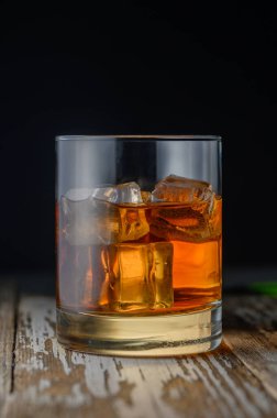 Altın bir içecek ve buzla dolu bir bardak, yumuşak ışığın altında parlayan, ahşap bir masanın üzerinde durur..