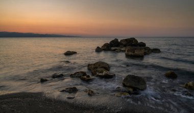Dalgalar günbatımının yumuşak renklerini yansıtan huzurlu bir alacakaranlık gökyüzünün altında kayalık kıyılarda nazikçe tur atar..