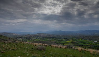 Canlı yeşil tarlaların ve kasvetli gökyüzünün altında uzak dağların üzerinde dramatik bulutlar toplanır..