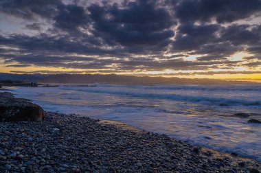 Dalgalar alacakaranlıkta dalgalanan canlı bulutlar gibi kayalık bir sahile çarpar, nefes kesici bir kıyı başyapıtı çizer..