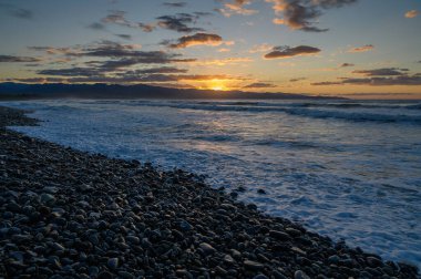 Altın güneş ufukta beliriyor, çakıllı kumsalları aydınlatırken dalgalar alacakaranlıkta sahili sarıyor..