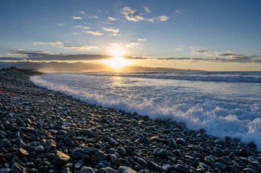 Altın ışınlar kıyı boyunca çakıl taşlarını aydınlatırken dalgalar alacakaranlıkta canlı bir gökyüzü altında yavaşça çarpışıyor..
