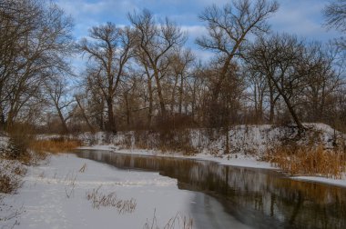 Durgun bir nehir, karla kaplı kıyılar ve yapraksız ağaçlarla çerçevelenmiş bir kış manzarası boyunca akar, sükuneti çağrıştırır..