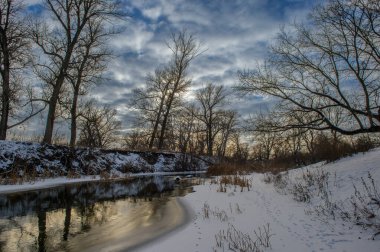 Bulutlu bir gökyüzünün altında, sakin bir nehir karla kaplı ağaçlar ve sessiz çevrelerle çevrili solmuş ışığı yansıtıyor..