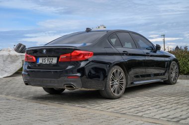 Gaziveren Kıbrıs Rum Kesimi -28.02.2025 Alacakaranlıkta kentsel manzarayla çevrili asfalt bir yüzeye park etmiş parlak siyah bir sedan duruyor.