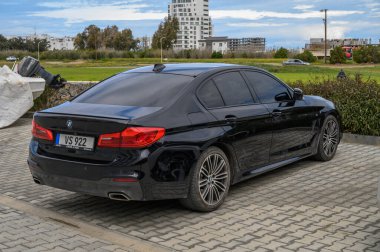 Gaziveren Kıbrıs Rum Kesimi -28.02.2025 Parlak siyah bir sedan açık bir gökyüzünün altında çağdaş binaların bulunduğu yeşil bir tarlanın yanına park edilmiştir..