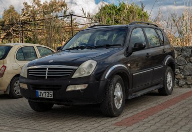 Gaziveren Kıbrıs Rum Kesimi - 28.02.2025 Siyah bir SUV, bulutlarla dolu bir gökyüzünün altında, yeşil bir arabanın yanında sürücüsünü bekliyor..