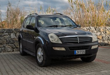 Gaziveren Kıbrıs Rum Kesimi -28.02.2025 Klasik siyah bir SUV engebeli bir taş duvarın ve bulutlu gökyüzünün altındaki uzun yeşilliğin yanında asfalt bir alana park edilmiştir..