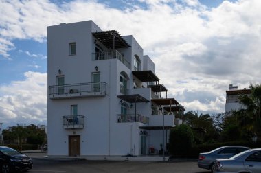 Modern bir bina, yeşil alanlarla çevrili, balkonları ve parlak bir gökyüzünün altında park edilmiş arabalarıyla ayakta duruyor..