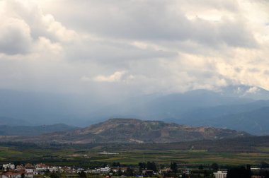 Geniş bir görünüm dalgalı dağları, kırsal bir köyü ve yağmuru tehdit eden karanlık bulutları gözler önüne seriyor..