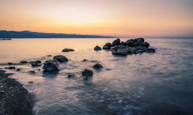Yumuşak dalgalar, nefes kesen bir gün batımının altında kayalık kıyılara doğru süzülüyor. Ufuk boyunca sıcak renkler döküyor..