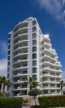 Zarif beyaz bir bina, canlı mavi bir gökyüzünün altında, mülkü çevreleyen palmiye ağaçları olan şık balkonlarla övünür..