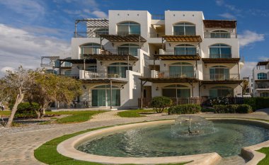 Sersemletici mimari beyaz duvarlar ve birden fazla balkon sergiliyor, canlı yeşillikler arasında sakin bir çeşmeyi çerçeveliyor.