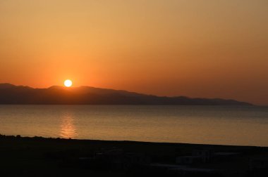 Güneş sakin kıyı şeridi boyunca uzak dağların ardında batarken altın ışınlar ufku aydınlatıyor..
