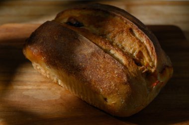 Taze pişmiş ekmeğin altın kahverengi kabuğu ahşap bir kesme tahtasının üzerinde oturur, güneş ışığı onun dokusunu aydınlatır..