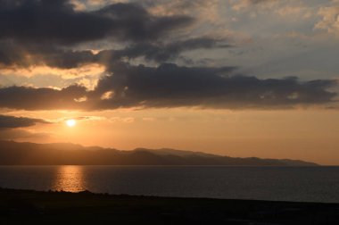 Gün batımının altın ışıkları bulutların altında yükselen uzak dağlar gibi sakin suları yansıtır..