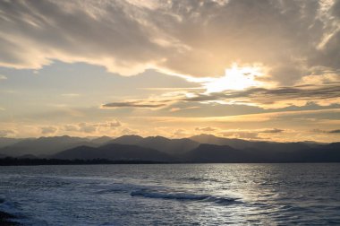 Sönük dalgalar yavaşça kıyıya vurur canlı bir günbatımı uzak dağların üzerinde sıcak renklerle gökyüzünü boyarken.