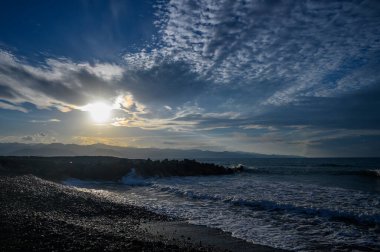 Güneş batarken, dalgalar yavaşça çakıl taşlı bir kıyıya çarpar. Gökyüzünü mavi ve altın renkleriyle boyarlar..