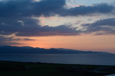 Gün batımında gökyüzünde harmanlanan canlı renkler sakin denize büyüleyici yansımalar yapıyor. Dağların gölgeleri, alacakaranlığın güzelliğini yansıtan sakin bir zemin oluşturur..