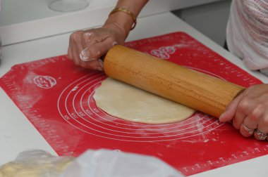 Modern bir mutfakta hamuru parlak kırmızı bir hasırın üzerine ustalıkla seren bir insan hamur işi yapmaya hazırlanıyor. Sıcak ortam aşçılık yaratıcılığını davet ediyor, yemek meraklıları için mükemmel..