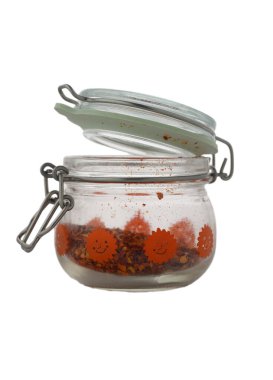 Canlı cam bir kavanoz neşeli bir tasarımla açık durur ve ezilmiş kırmızı biber tanelerinin içeriğini gösterir. Sıcak öğleden sonra güneşi zengin renkleri vurguluyor, mutfak atmosferine sıcaklık katıyor..