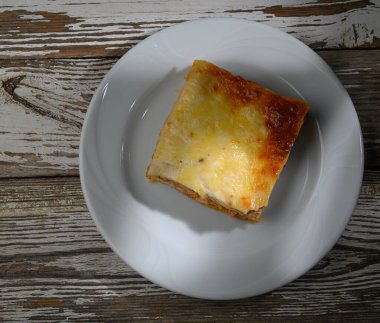 Lezzetli bir kare fırınlanmış lazanya temiz beyaz bir tabağın üzerinde duruyor, makarna, peynir ve zengin et sosu tabaklarını sergiliyor, hepsi büyüleyici bir ahşap yüzeye karşı..