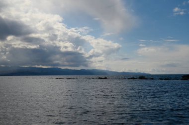 Dalgalanan su ufuk boyunca uzanıyor, bulutlarla dolu bulutlu bir gökyüzü ile karşılaşıyor. Uzak dağlar, göl kenarında huzurlu ama dinamik bir atmosfer yaratıyor..