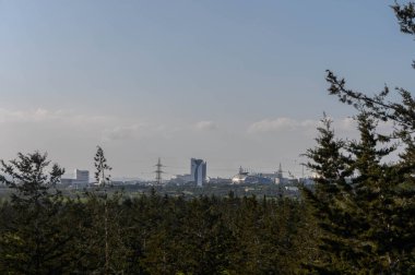 Parlak mavi bir gökyüzünün altında güneşlenen geniş bir şehir manzarasını çevreleyen canlı ağaçlar..